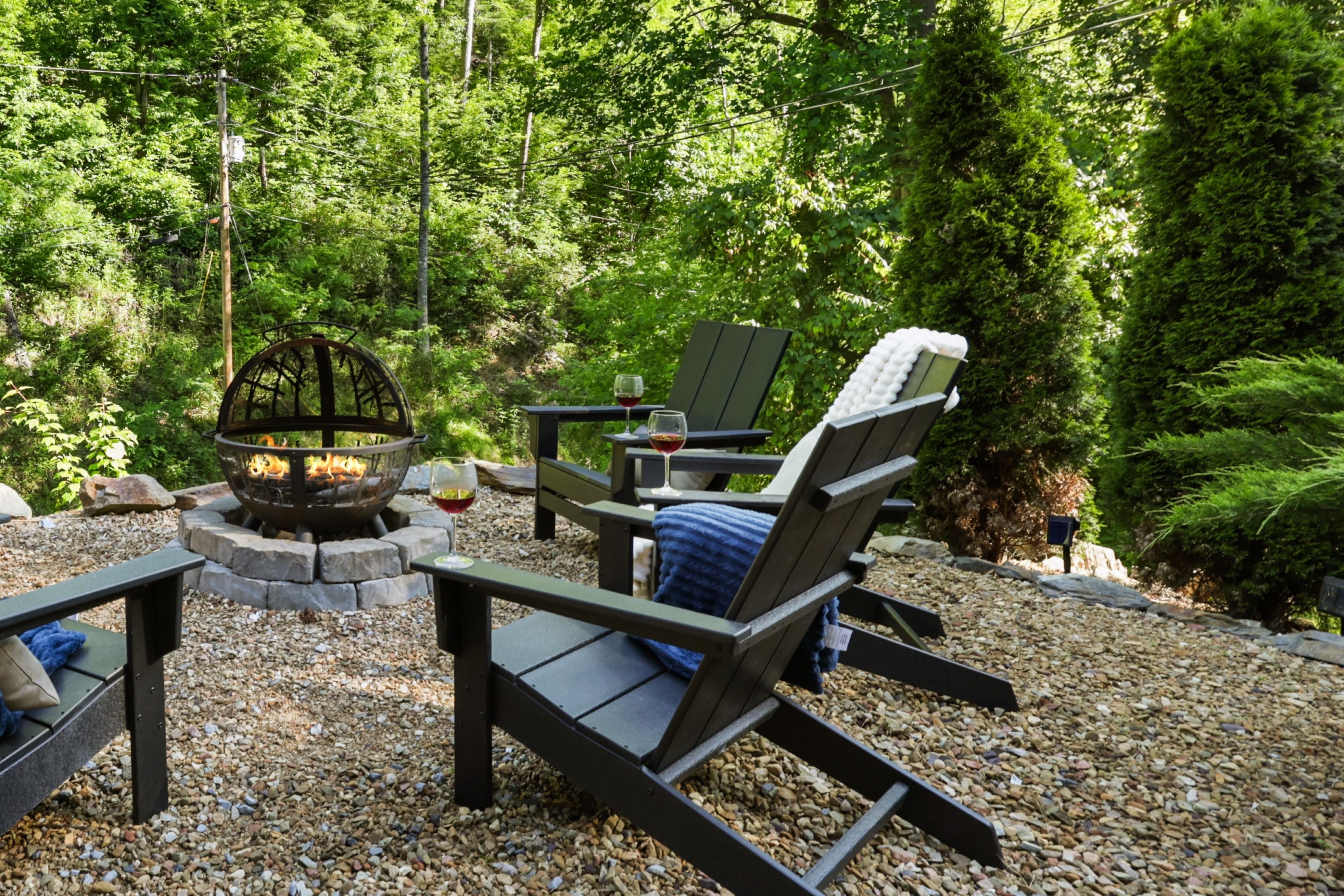 Outdoor setting with Adirondack chairs around a fire pit in a forested area.