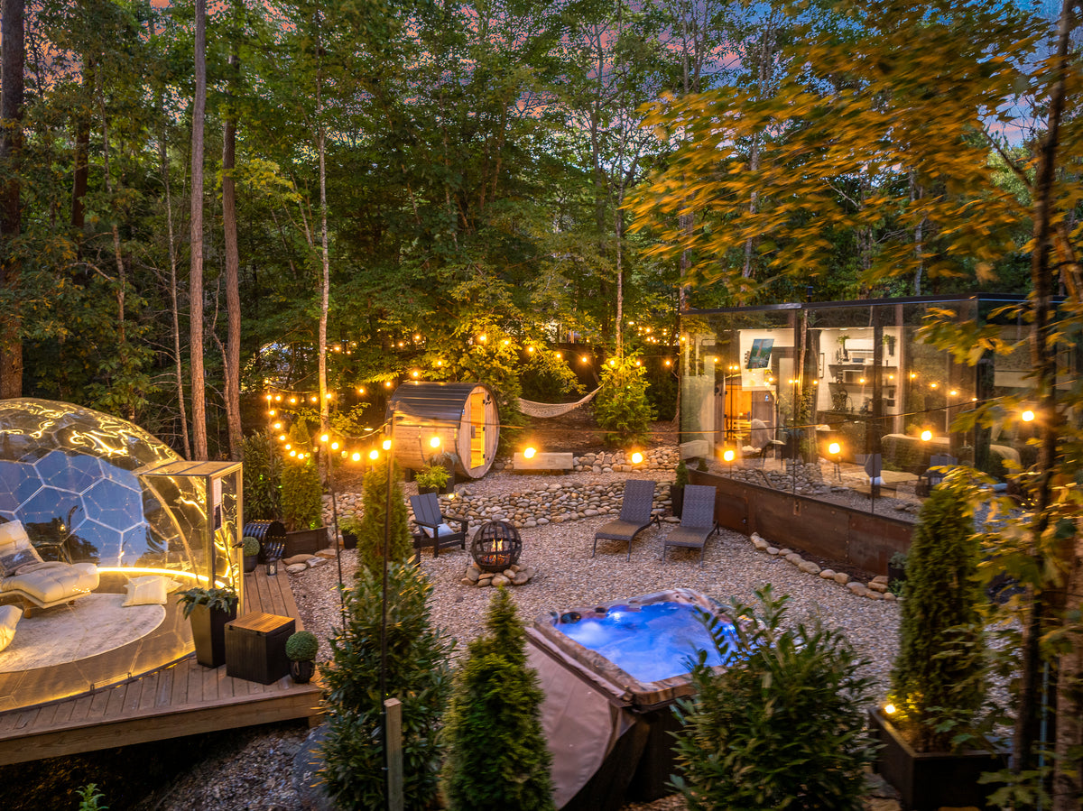 A luxury Villa Sol by Evefrwild Stays outdoor retreat backyard at dusk featuring a glass geodome tent, a modern glass cabin, a small illuminated hot tub, a hammock, and a gravel lounge area surrounded by warm string lights in a forest setting.